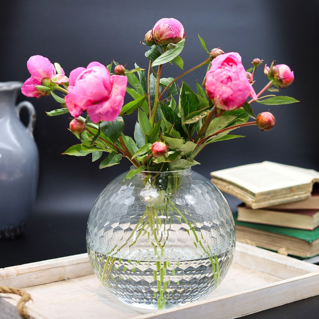 Clear glass vase with pink flowers on a wooden surface with books in the background