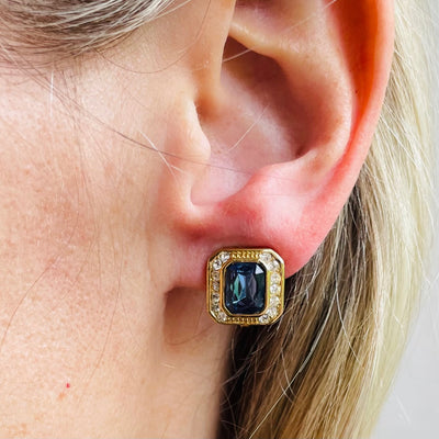 A matching pair of vintage Christian Dior clip-on earrings against a plain white background. The earrings are thickly gold-plated and feature a deep emerald-cut sapphire blue crystal stone in the center, encircled by a precise halo of small, pave-set clear round rhinestones.