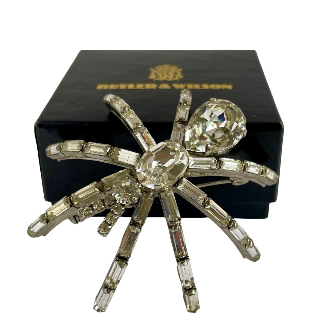 A very large 1980s Butler & Wilson spider brooch with clear faceted crystals on a silver-tone metal base, displayed in front of a black Butler & Wilson box on a white background.