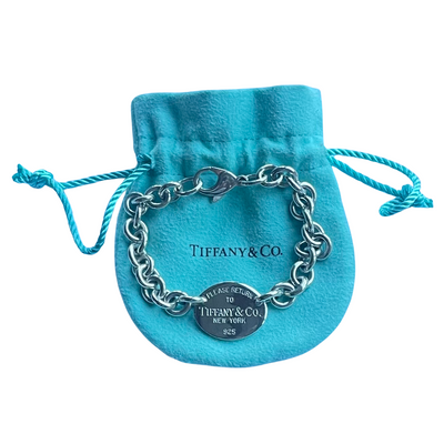 A silver chain bracelet with a clasp and a round signature tag, displayed on a  blue velvet drawstring pouch with 'Tiffany & Co.' printed on it, against a plain white background.