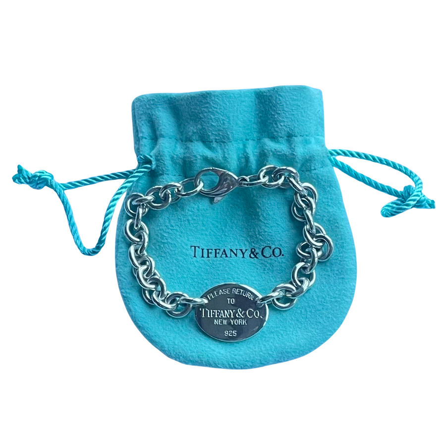 A silver chain bracelet with a clasp and a round signature tag, displayed on a  blue velvet drawstring pouch with 'Tiffany & Co.' printed on it, against a plain white background.