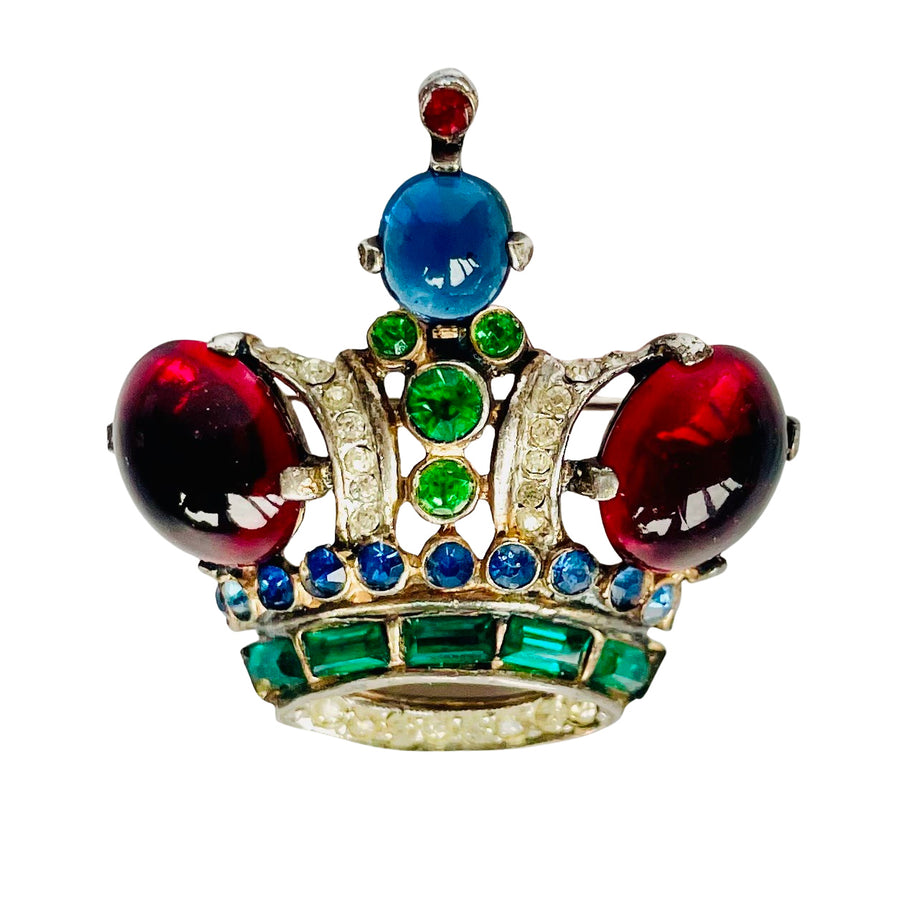 Front view of a vintage 1944 Crown Trifari sterling silver crown brooch designed by Alfred Philippe. The brooch features a vibrant regal color palette including two large, high-domed red glass cabochons on either side, a central sapphire blue glass cabochon at the peak, and a row of emerald-green crystal baguettes at the base. The silver arches and base are intricately accented with small clear and blue rhinestones against a clean white background.