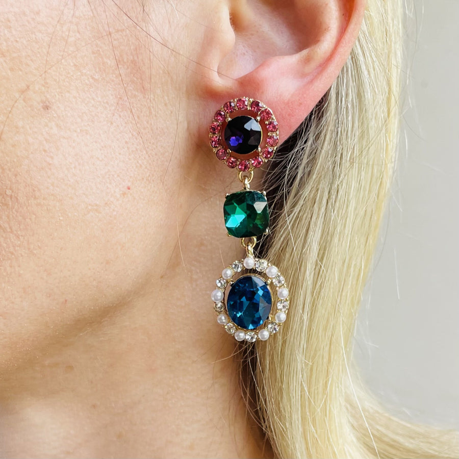 Close up on model's ear of three-tiered gold tone Monet drop earrings featuring purple, green, and royal blue crystals with faux pearls.