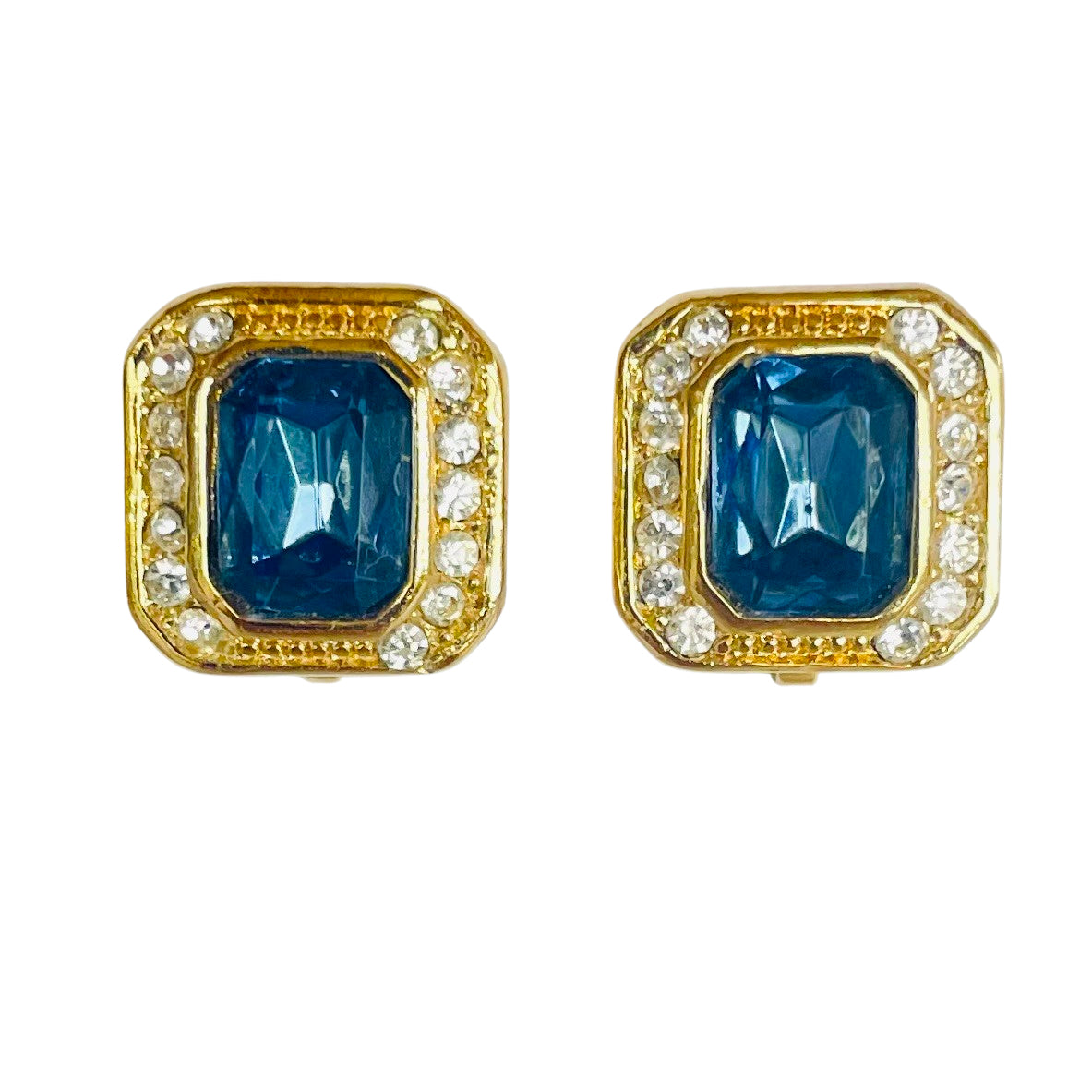 A matching pair of vintage Christian Dior clip-on earrings against a plain white background. The earrings are thickly gold-plated and feature a deep emerald-cut sapphire blue crystal stone in the center, encircled by a precise halo of small, pave-set clear round rhinestones.
