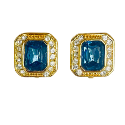 A matching pair of vintage Christian Dior clip-on earrings against a plain white background. The earrings are thickly gold-plated and feature a deep emerald-cut sapphire blue crystal stone in the center, encircled by a precise halo of small, pave-set clear round rhinestones.