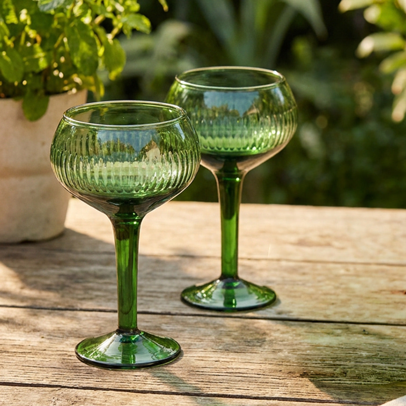 A pair of green glass coupe-style goblets or martini glasses with a ribbed textured bowl and a slender stem displayed on a rustic table with blurred background.