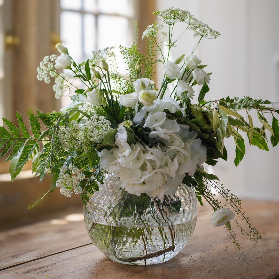 Clear glass vase with a bouquet of white and green flowers on a wooden surface.