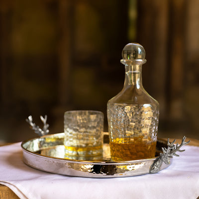 A Culinary Concepts round silver metal tray with a hammered texture and decorative stag handles on both sides, shown on a table with decanter and glass