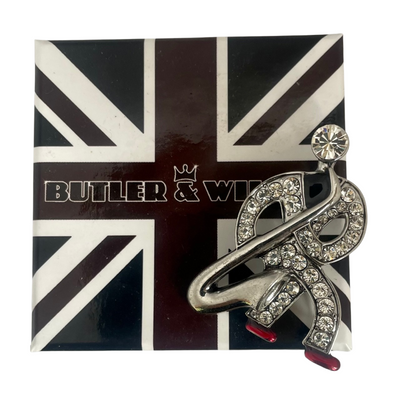 Front view of a silver-tone Butler & Wilson Art Deco style saxophonist brooch against a white background, featuring clear rhinestones and red enamel shoes on signature Butler & Wilson black, white and red box.
