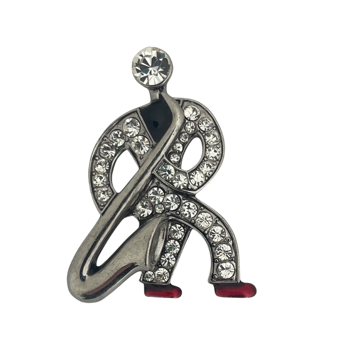Front view of a silver-tone Butler & Wilson Art Deco style saxophonist brooch against a white background, featuring clear crystals and red enamel shoes.
