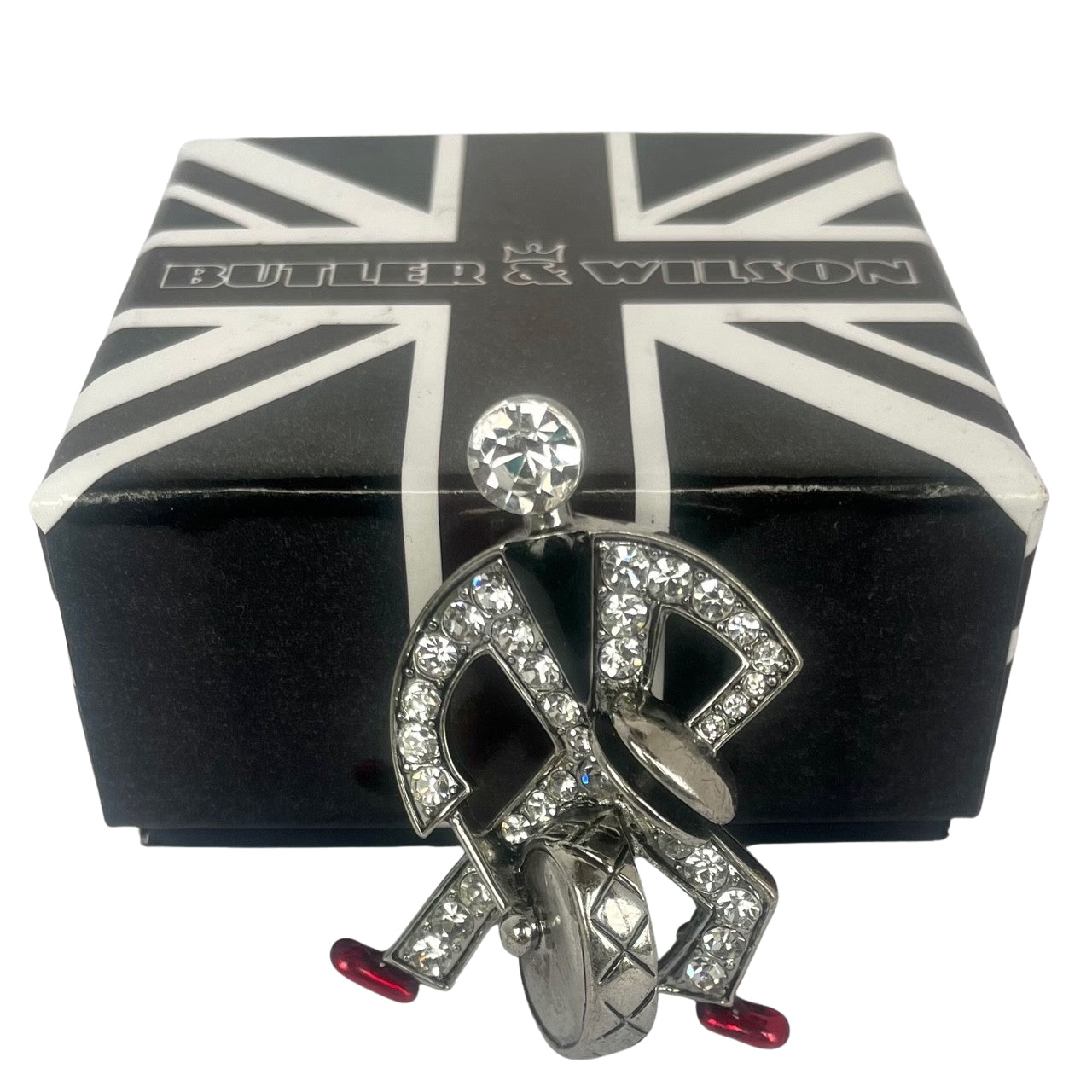 Front view of a silver-tone Butler & Wilson Art Deco style jazz drummer brooch, adorned with clear crystal rhinestones, black enamel, and red enamel shoes, propped vertically against its black and white Union Jack logo box