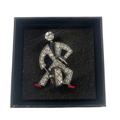 Front view of a silver-tone Butler & Wilson Art Deco style jazz drummer brooch, adorned with clear crystal rhinestones, black enamel, and red enamel shoes, laying in its black and white Union Jack logo box