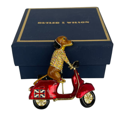 A Butler & Wilson enamel pin of a brown dachshund wearing a rhinestone-embellished sweater and riding a red scooter with gold trim, set against a blue branded Butler & Wilson box.