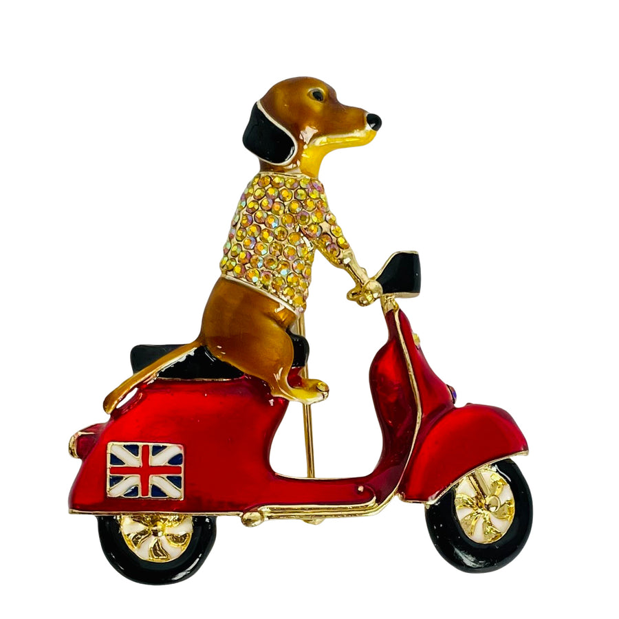 A Butler & Wilson enamel pin of a brown dachshund wearing a rhinestone-embellished sweater and riding a red scooter with gold trim, set against a plain white background.