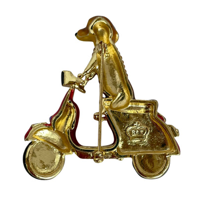 reverse of  Butler & Wilson enamel pin of a brown dachshund wearing a rhinestone-embellished sweater and riding a red scooter with gold trim, set against a plain white background.