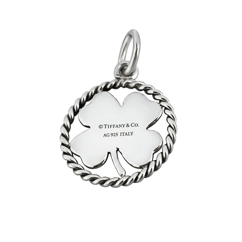 Back view of a polished silver four-leaf clover charm by Tiffany and Co. The clover leaf is enclosed in a circular twisted wire rope border and features a silver jump ring at the top. The centre is engraved with the text copyright Tiffany and Co, AG 925 ITALY.
