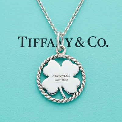 A polished silver four-leaf clover pendant by Tiffany and Co shown on Tiffany & Co box. The clover leaf is enclosed in a circular twisted wire rope border and features a silver jump ring at the top. The centre is engraved with the text copyright Tiffany and Co, AG 925 ITALY.