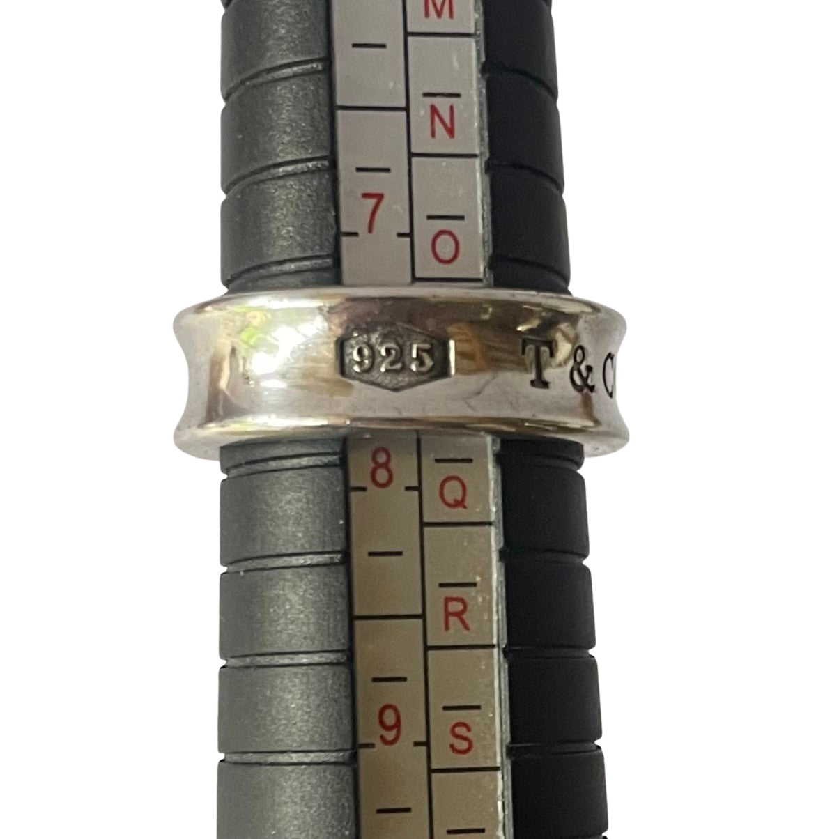 A silver Tiffany & Co. ring being measured on a black metal mandrel ring sizer. The ring sits between sizes 7 and 8, showing the "925" and partial "T & CO" engravings on its outer band.