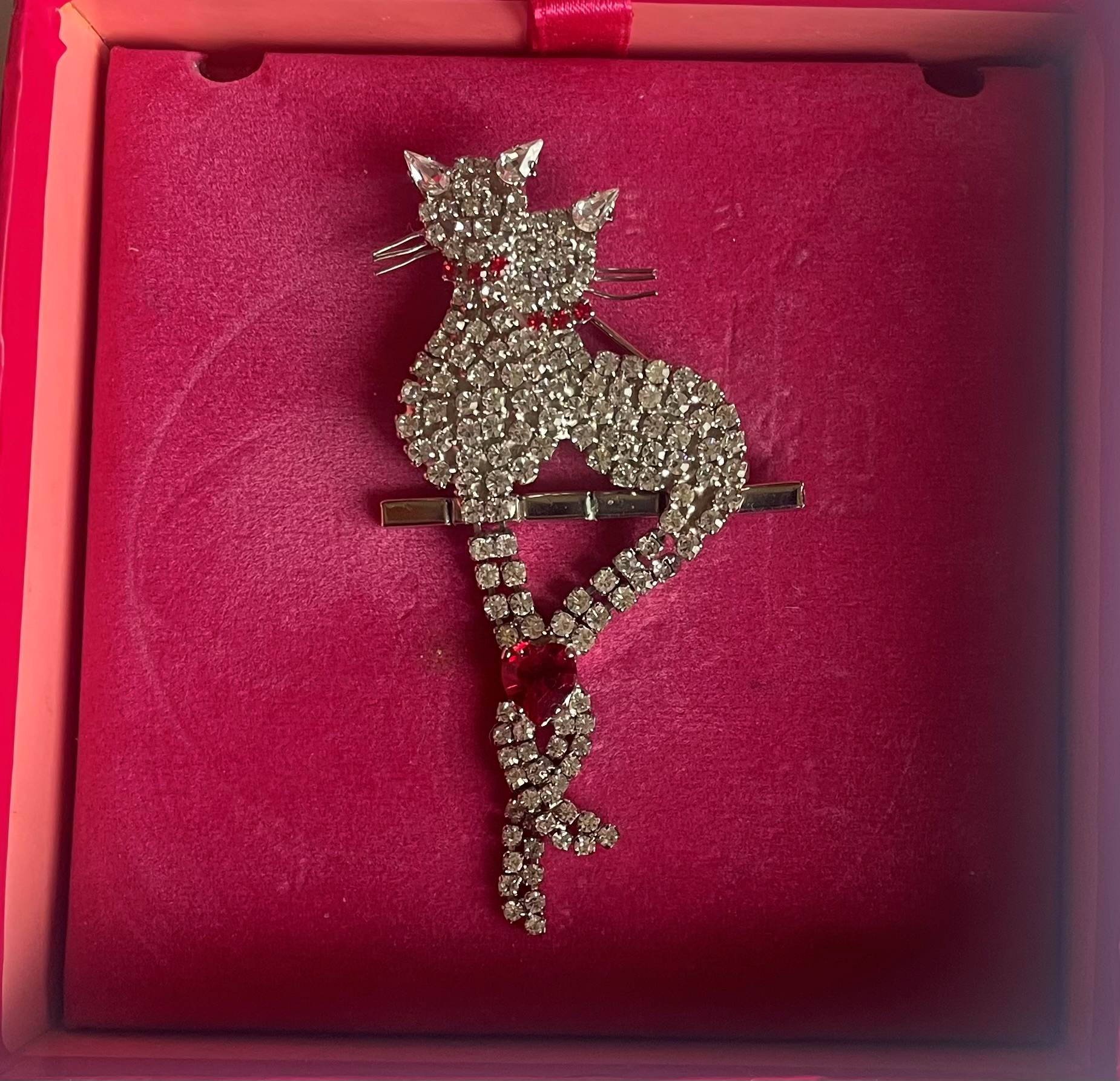 Butler & Wilson vintage crystal cat brooch featuring two pavé-set rhinestone cats perched on a bar, with their articulated tails intertwined and united by a vibrant ruby-red crystal heart in its original box.