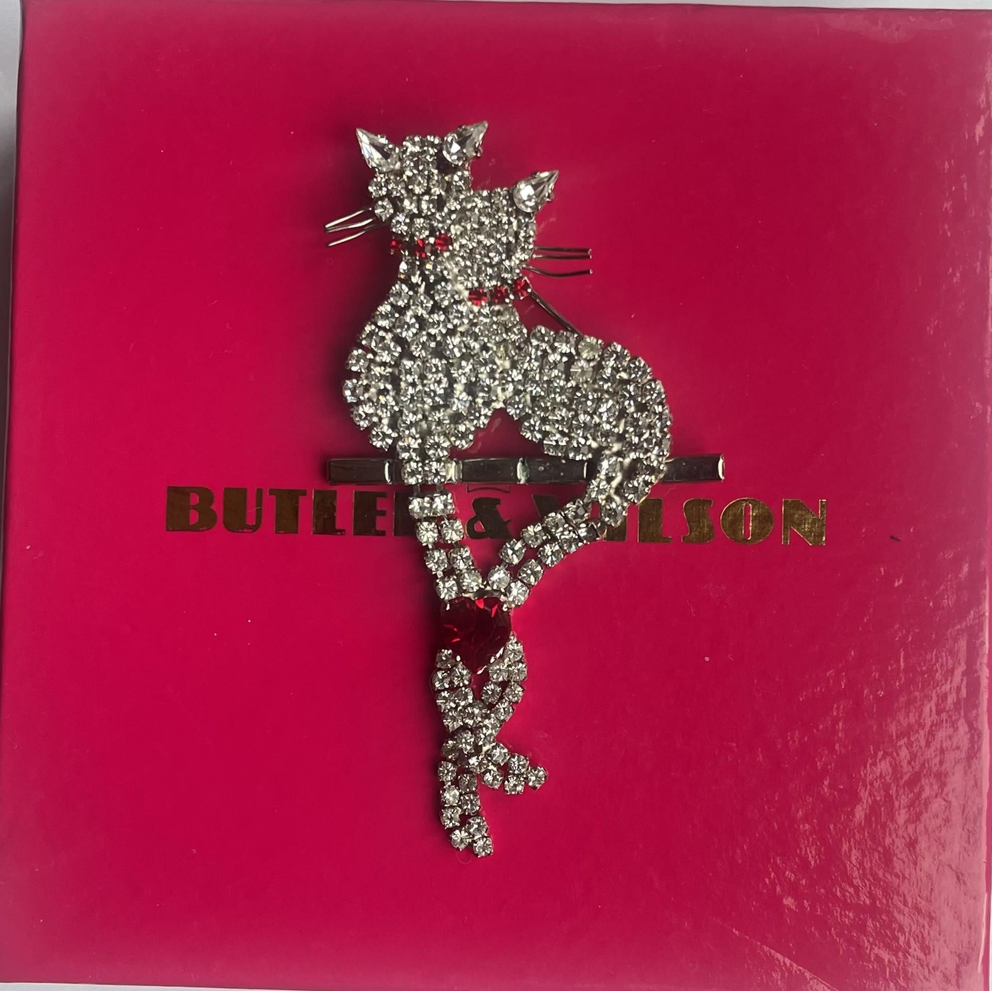 Butler & Wilson vintage crystal cat brooch featuring two pavé-set rhinestone cats perched on a bar, with their articulated tails intertwined and united by a vibrant ruby-red crystal heart on its original box.
