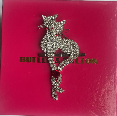 Butler & Wilson vintage crystal cat brooch featuring two pavé-set rhinestone cats perched on a bar, with their articulated tails intertwined and united by a vibrant ruby-red crystal heart on its original box.