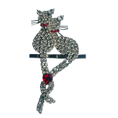 Butler & Wilson vintage crystal cat brooch featuring two pavé-set rhinestone cats perched on a bar, with their articulated tails intertwined and united by a vibrant ruby-red crystal heart on a white background.