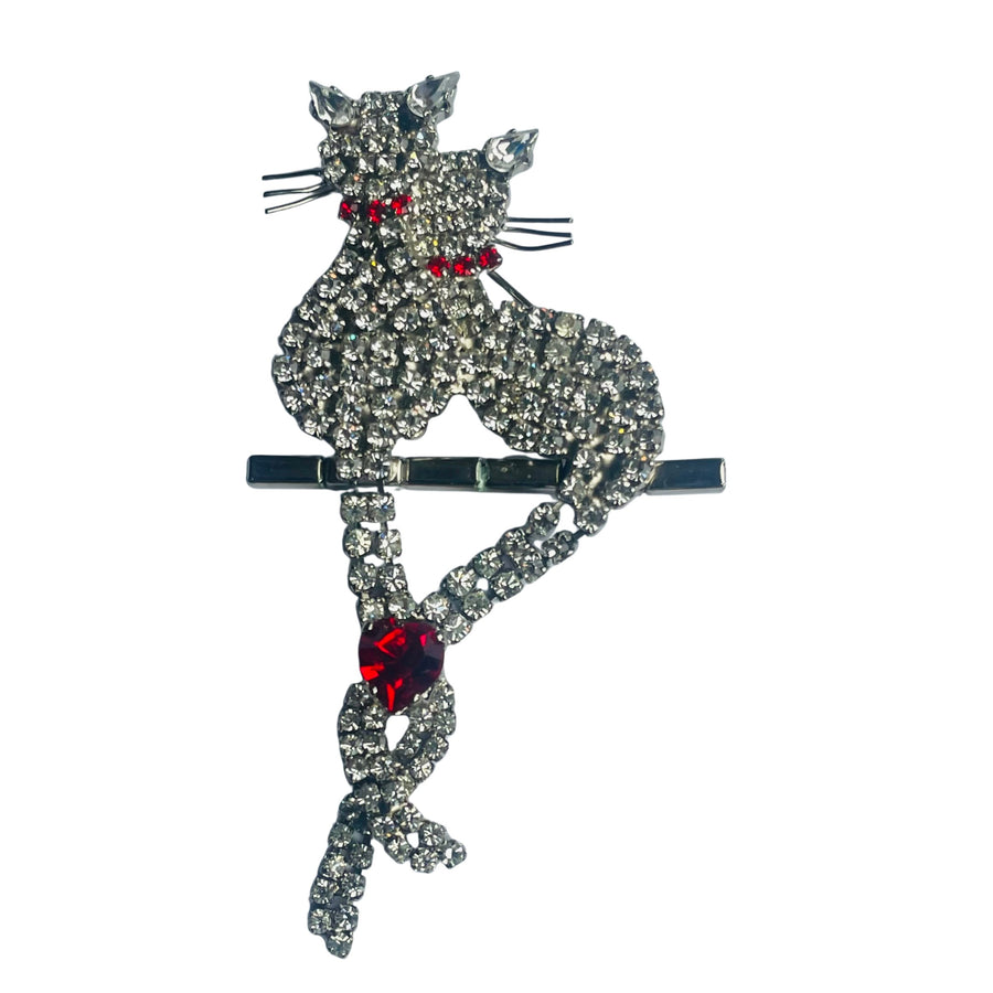 Butler & Wilson vintage crystal cat brooch featuring two pavé-set rhinestone cats perched on a bar, with their articulated tails intertwined and united by a vibrant ruby-red crystal heart on a white background.