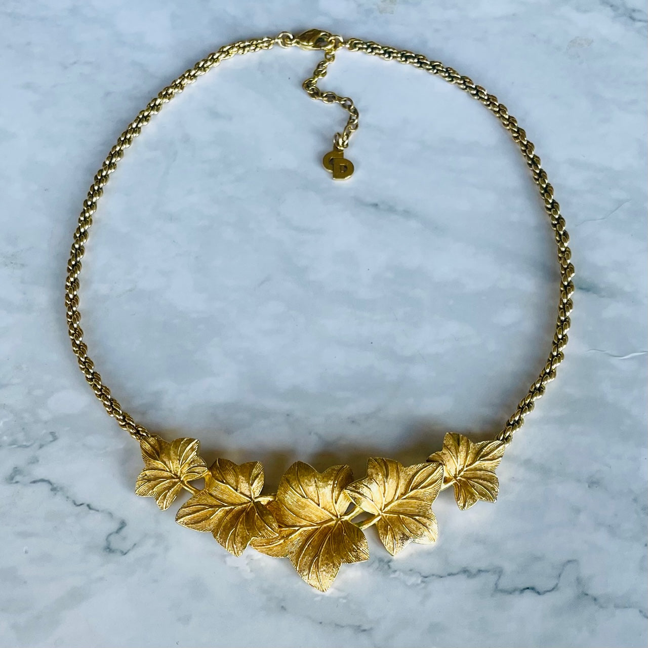 A full flat-lay view of a vintage 1980s Christian Dior gold-plated necklace arranged in a neat circle on a white and grey veined marble surface. The lower portion showcases the five detailed, textured ivy leaves forming a curved pendant focal point, which connects to a twisted rope chain. At the top centre the lobster claw clasp is fastened to an adjustable extender chain that finishes with a small, circular hanging "CD" logo charm.