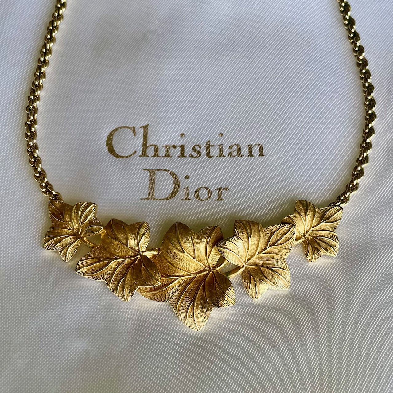 Close-up shot of a vintage 1980s Christian Dior gold-tone necklace featuring five detailed ivy leaves with a textured, brushed finish. The necklace is laid horizontally across a cream-colored satin pouch, with the words "Christian Dior" clearly printed in an elegant serif font directly above the leaves.