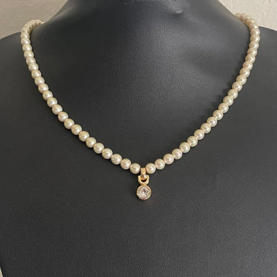 A vintage Christian Dior single-strand cream faux pearl necklace displayed on a black neck mannequin. The necklace features a central gold-plated pendant drop hanging from a distinct horseshoe-shaped bail, holding a brilliant, bezel-set round clear solitaire crystal.