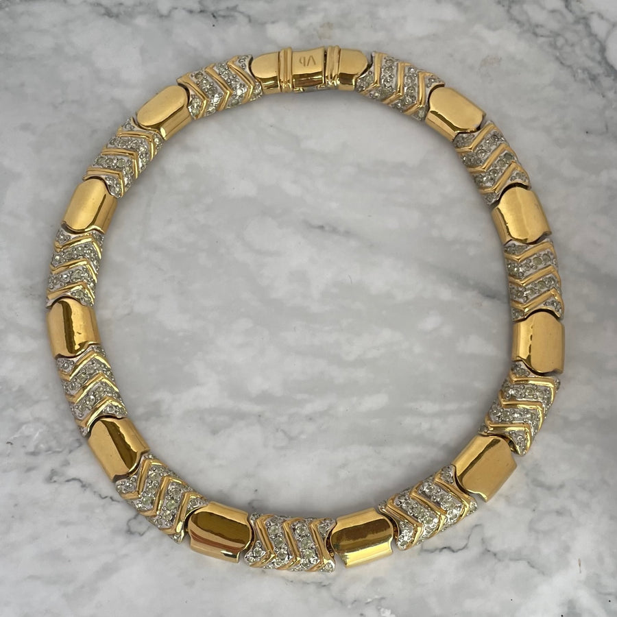 intage 1980s Vogue Bijoux heavy collar choker necklace arranged in a circle on a gray veined marble surface. The necklace features a chunky, repeating architectural pattern of alternating high-polished, smooth gold-tone rectangular blocks and textured chevron-shaped links encrusted with clear pavé crystals. At the top of the circle, the seamless integrated clasp is visibly stamped with the initials "Vb"