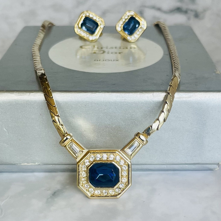 A vintage 1980s Christian Dior gold-plated demi-parure set featuring a flat herringbone chain necklace with a central octagonal pendant and matching square clip-on earrings. Both pieces showcase an emerald-cut sapphire blue crystal stone framed by clear round rhinestones, displayed in front of a grey vintage "Christian Dior BIJOUX" jewelry box.