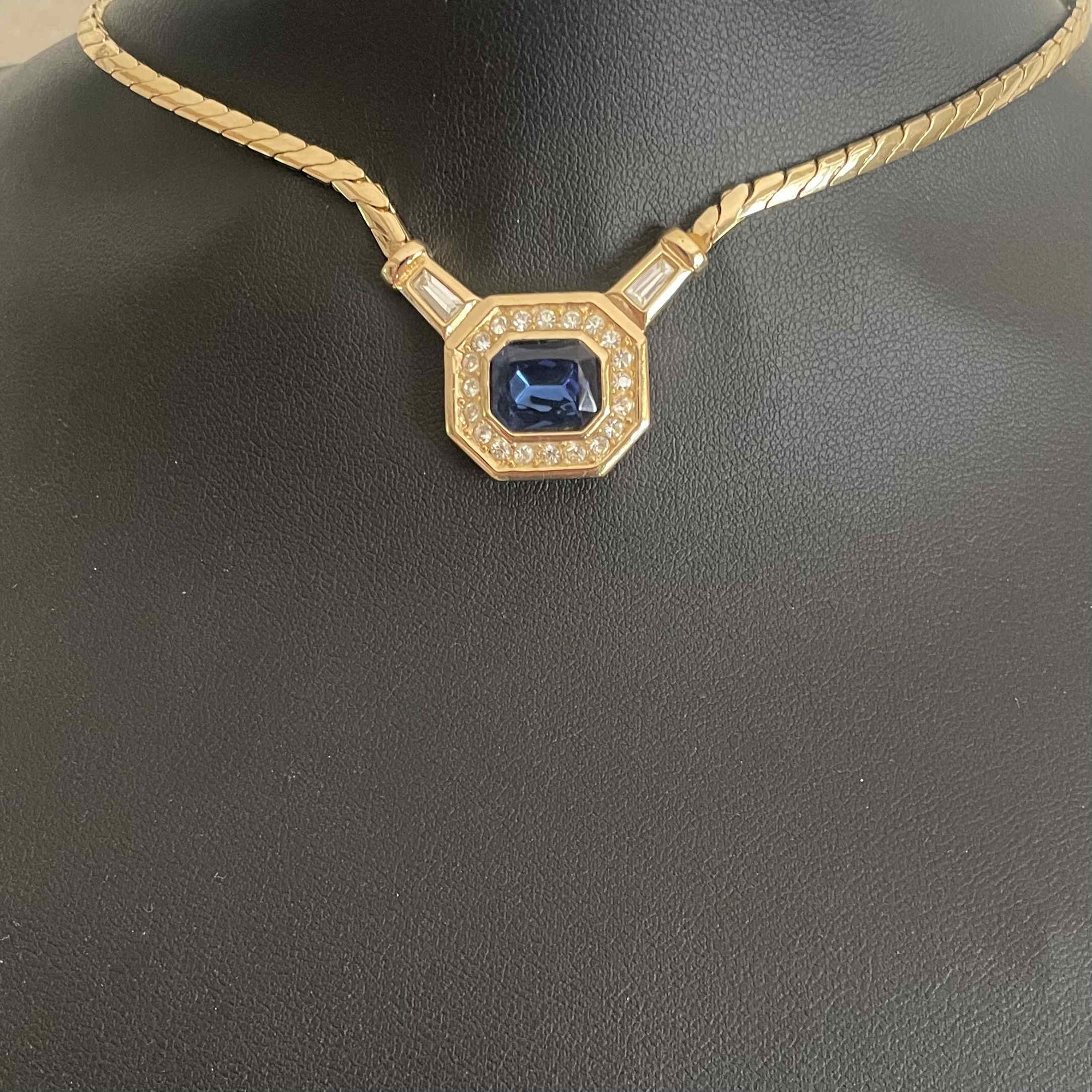 Close-up of a vintage Christian Dior gold-plated necklace displayed on a black mannequin bust. The necklace features a fluid herringbone chain leading to an octagonal pendant set with a large, step-cut sapphire-blue central crystal, surrounded by a border of brilliant-cut clear rhinestones. Two clear baguette rhinestones accent the chain connections.